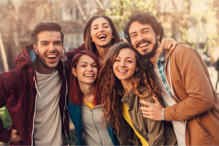 Five friends smiling with arms around each other outdoors, showing supportive community and connection.