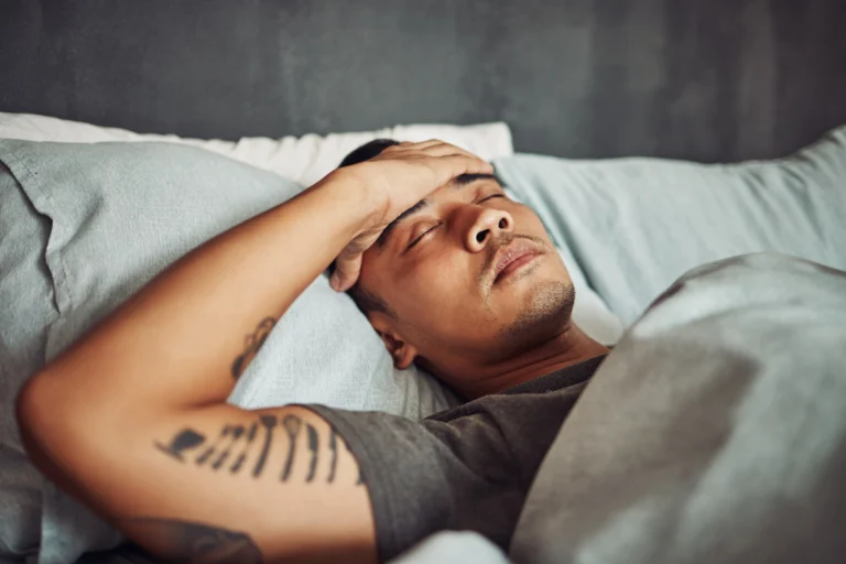 Man lying in bed with his hand on his forehead, appearing unwell with symptoms that can occur during early alcohol withdrawal.