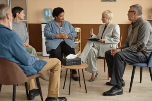 Facilitator leading a seated circle during an addiction group therapy session, with participants listening and one speaking.